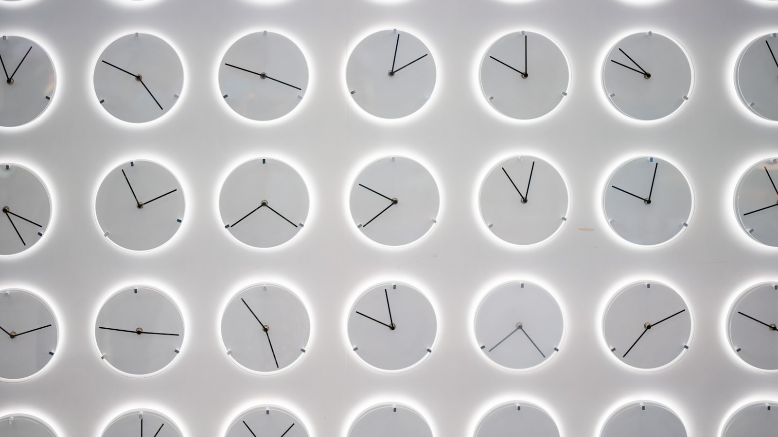 A brightly lit white wall covered in rows of identical minimalist white clock faces, each ringed by a soft backlit halo and each showing a different time