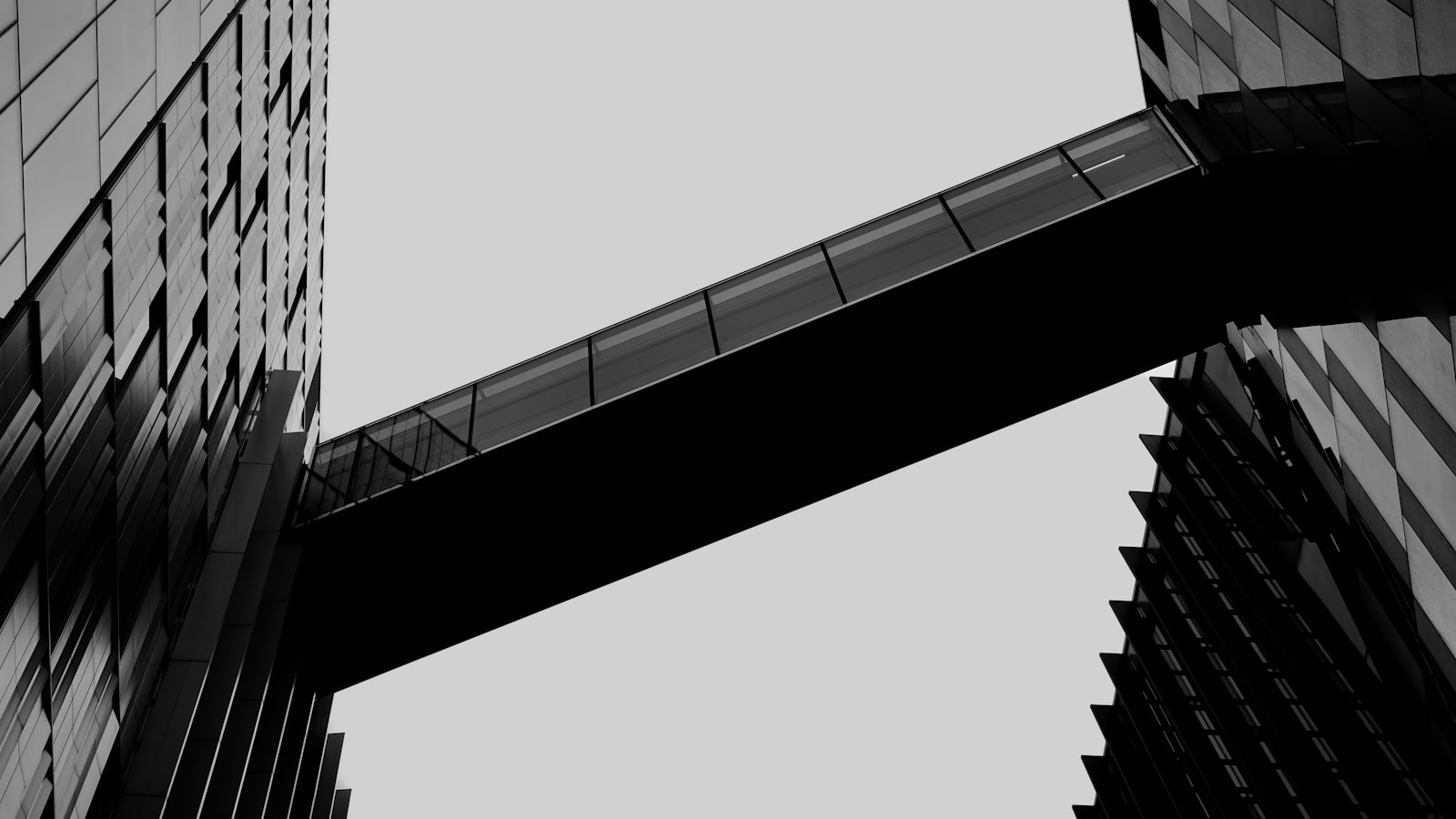 Low-angle black and white photograph of a glazed skybridge running diagonally across a pale sky to join two dark glass office towers