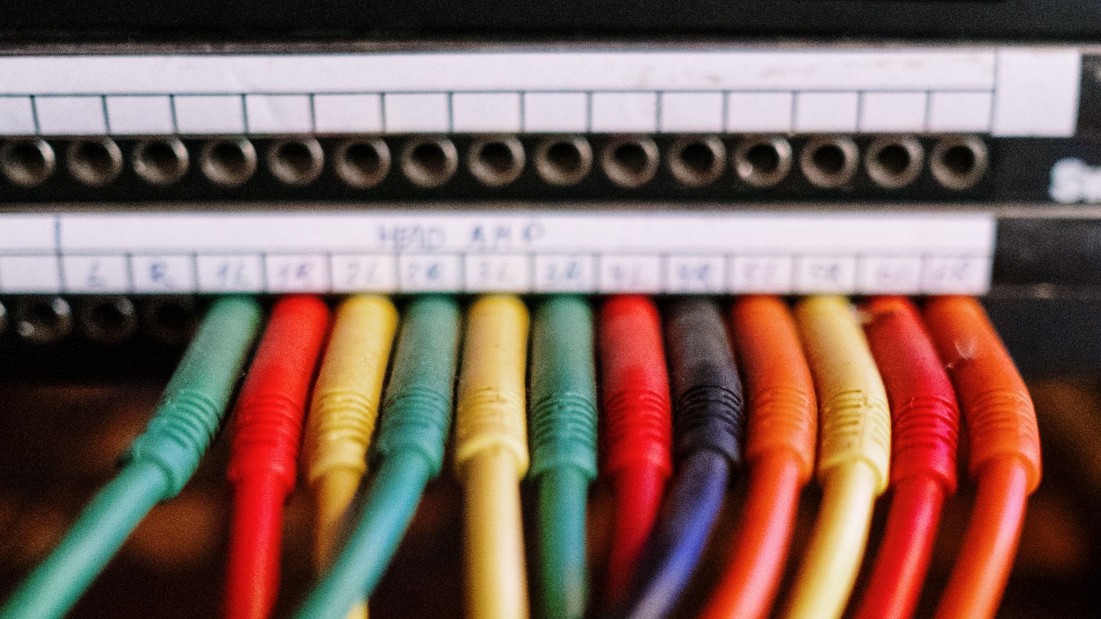 Close-up of a studio patch bay: two rows of hand-labelled jack ports above a row of green, red, yellow, orange and dark blue patch cables plugged in side by side and sweeping down out of frame together